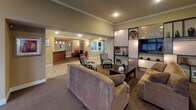 Located at 32905 19th Pl S in Federal Way, Washington, this single-story community clubhouse offers a thoughtfully designed space for both relaxation and activity. The welcoming lounge area serves as the heart of the facility, featuring a classic fireplace framed by a dark surround, built-in display shelving, a large flat-screen television, and comfortable seating arranged for conversation and leisure. Warm, neutral tones and recessed lighting create an inviting ambiance throughout. Adjacent to the lounge, a reception area with a wood-front counter provides a functional point of entry. A separate dining and gathering room highlights soaring vaulted ceilings with a dramatic arched window that floods the space with natural light and views of lush greenery. This room includes a round dining table with seating and a built-in kitchenette with rich wood cabinetry, a sink, and counter space — ideal for community events. The fitness center is generously sized, featuring vaulted ceilings, large windows overlooking the outdoors, full-length mirrors, and a comprehensive array of equipment including a stationary bike, elliptical, cable machine, and weight bench, all positioned on protective floor mats.