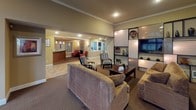 Located at 32905 19th Pl S in Federal Way, Washington, this single-story community clubhouse offers a thoughtfully designed space for both relaxation and activity. The welcoming lounge area serves as the heart of the facility, featuring a classic fireplace framed by a dark surround, built-in display shelving, a large flat-screen television, and comfortable seating arranged for conversation and leisure. Warm, neutral tones and recessed lighting create an inviting ambiance throughout. Adjacent to the lounge, a reception area with a wood-front counter provides a functional point of entry. A separate dining and gathering room highlights soaring vaulted ceilings with a dramatic arched window that floods the space with natural light and views of lush greenery. This room includes a round dining table with seating and a built-in kitchenette with rich wood cabinetry, a sink, and counter space — ideal for community events. The fitness center is generously sized, featuring vaulted ceilings, large windows overlooking the outdoors, full-length mirrors, and a comprehensive array of equipment including a stationary bike, elliptical, cable machine, and weight bench, all positioned on protective floor mats.