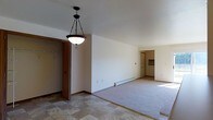 This single-floor home presents an open-concept layout that integrates the living, dining, and kitchen areas into one continuous space. The living area is carpeted and accommodates a comfortable seating arrangement, including cushioned armchairs draped in light fabric and a small side table accented with decorative flowers. A textured area rug anchors the seating zone. The ceiling features a drop-tile design throughout, maintaining a consistent, low-profile look across the space.

Transitioning from the living area, the kitchen is visible through an open passageway and is equipped with medium-toned wood cabinetry on both upper and lower sections, a white electric range with overhead ventilation, a dishwasher, a full-size refrigerator, and a microwave. The countertops provide modest workspace, and under-cabinet lighting adds functional illumination. A double sink is positioned along the counter near the window area.

Adjacent to the kitchen, a small dining area accommodates a table and upholstered chairs, with a ceiling fan overhead providing air circulation. The overall ambiance is modest and functional, with a warm, lived-in character throughout.