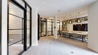 Located at 195 River Grove Way in West Palm Beach, Florida, this single-floor business center offers a thoughtfully designed workspace defined by a sleek, contemporary aesthetic. The space is anchored by a spacious conference room outfitted with a long, neutral-toned table, ergonomic rolling chairs, and a large wall-mounted display screen. Built-in open shelving lines the walls, and a pair of striking industrial-style pendant lights hang overhead, adding a refined, modern touch. The flooring throughout features a chevron-patterned light tile that contributes to the clean, professional atmosphere. A defining architectural feature of the space is the extensive use of black-framed glass partition walls and doors, which divide the area into distinct zones — including private meeting rooms and a lounge area — while maintaining a sense of openness and transparency. One area opens to views of lush palm trees, a pool, and the surrounding outdoor grounds through floor-to-ceiling black-framed windows, bringing natural light and a tropical ambiance into the interior. A cozy seating nook with a rounded upholstered chair is positioned near these windows alongside a printer station, blending comfort with function. The overall design balances professionalism with warmth, making it a versatile and well-appointed environment for work and collaboration.