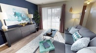 This single-floor space offers a thoughtfully designed interior that balances comfort and contemporary style. The living area serves as the centerpiece of the home, anchored by a bold navy accent wall that adds depth and character to the room. Large sliding glass doors with vertical blinds and elegant mauve draping panels allow an abundance of natural light to pour in, creating a warm and inviting atmosphere while offering a view of the outdoors.

The flooring features wood-look planks that lend a clean, modern aesthetic throughout the space. A stylish area rug defines the seating zone, where a plush sectional sofa in cool grey tones pairs seamlessly with coordinating accent pillows. A sleek dark-toned coffee table and a contrasting teal tray add pops of color and visual interest to the arrangement.

Along the accent wall, a low-profile grey dresser provides ample storage while serving as a display surface for decorative items, including a statement table lamp and a vibrant abstract canvas print. Lush indoor greenery introduces a refreshing natural element to the space. Overall, the space exudes a relaxed yet polished ambiance, making it ideal for both everyday living and entertaining.