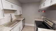 This single-story unit in North Richland Hills, Texas, offers a well-designed layout with a mix of modern updates and functional living spaces. The living room serves as the centerpiece of the home, showcasing dramatic vaulted ceilings with a ceiling fan, light wood-tone flooring, and a brick-surround fireplace flanked by built-in shelving. Natural light filters through window blinds, enhancing the open, airy feel of the space.

The kitchen is efficiently arranged in a galley-style configuration, featuring white shaker cabinetry with brushed nickel hardware, granite-look countertops, a white subway tile backsplash, and stainless steel appliances including a dishwasher and electric range. Light wood-tone flooring continues throughout this area.

The bedroom is carpeted and includes a ceiling fan with light fixture and a window with blinds providing natural light. Adjacent spaces include a hallway area with closet storage and wood-tone flooring, offering practical everyday functionality.

The bathroom is clean and bright, featuring a white vanity with shaker-style cabinetry, a tiled tub-shower combo, and wood-tone flooring. A dedicated laundry closet houses a full-size top-load washer and dryer, adding considerable convenience. Neutral gray walls and white trim are consistent throughout, giving the unit a cohesive, contemporary aesthetic.
