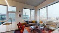 This single-floor urban residence showcases a thoughtfully designed open-concept layout that seamlessly connects the kitchen, dining, and living areas. The kitchen is outfitted with stainless steel appliances, including a refrigerator, dishwasher, and range, complemented by sleek white cabinetry that extends to the ceiling and a generous island with a built-in sink. Pendant lighting illuminates the island, which features bar seating for casual dining. Light wood-style flooring runs throughout the main living areas, contributing to a clean, contemporary aesthetic.

The living area benefits from an abundance of natural light streaming through large, floor-to-ceiling windows that frame sweeping city skyline views. Elevated ceilings enhance the sense of openness throughout the space.

The unit includes two bedrooms, each carpeted and featuring oversized windows with urban vistas. One bedroom offers views of a prominent city clock tower, while the other overlooks neighboring buildings.

The bathroom is finished with neutral tile flooring, a wide vanity with undermount sink, ample cabinetry, and a shower-tub combination. The overall design palette is modern and cohesive, combining white walls, clean lines, and contemporary fixtures throughout.