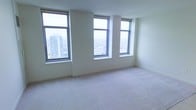 This high-rise residence offers a refined and airy living environment defined by its elevated position above the city skyline. The space is bathed in natural light, thanks to a series of large, floor-to-ceiling-style windows that frame sweeping panoramic views of the surrounding urban landscape, including neighboring high-rise buildings and the city stretching into the distance. The clean, neutral white walls create a bright and versatile backdrop, complementing the soft, wall-to-wall carpeting that adds warmth and comfort underfoot. The open layout provides a generous sense of space, ideal for flexible furniture arrangements and personalization. The windows are framed with sleek, dark trim that adds a modern contrast to the otherwise light and minimal interior palette. The elevated vantage point makes the cityscape a defining feature of the room, offering a dynamic and ever-changing view throughout the day. Overall, the space presents a clean, contemporary aesthetic with an emphasis on natural light, openness, and urban connectivity.