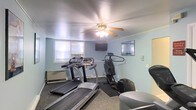 This single-floor fitness room provides a practical space for cardiovascular workouts and general exercise. The room is finished with light blue walls that create a calm, neutral atmosphere, complemented by carpeted flooring with rubber exercise mats placed beneath the equipment for added stability and protection. The space is equipped with multiple treadmills and a stair climber or elliptical machine, offering a variety of cardio options for users. A ceiling fan with an integrated light fixture is centrally mounted, helping to maintain airflow and comfort during workouts. Two wall-mounted flat-screen televisions are positioned for easy viewing from the equipment, enhancing the exercise experience. Natural light enters through windows along one wall, one of which houses a window air conditioning unit to help regulate the room's temperature. Baseboard heating along the perimeter ensures year-round comfort. The overall layout is straightforward and functional, making efficient use of the available space while providing the essential amenities needed for a well-rounded cardio fitness routine.
