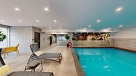 Located at 625 Milton in Montréal's vibrant Milton-Parc neighbourhood, this single-level amenity facility offers residents a comprehensive wellness experience. The space is divided into several distinct yet interconnected zones, each finished with a cohesive modern aesthetic featuring light stone-tile wall cladding, large-format porcelain flooring, and recessed lighting throughout.

The indoor swimming pool is a central highlight, featuring vivid turquoise water, lane markers, stainless steel entry rails, and a safety ring station. The pool area is flanked by a generous poolside lounge with reclining sun chairs and accent cushions, creating a resort-like atmosphere.

Adjacent to the pool, a well-lit fitness centre is visible through floor-to-ceiling glass partitions. It is equipped with cardio machines, resistance equipment, free weights, and exercise balls, all reflected in mirrored walls that enhance the sense of space.

A bright, windowed lounge area provides a welcoming social space furnished with vibrant yellow seating clusters and additional loungers, with large windows allowing ample natural light and views of the surrounding streetscape.

The facility also includes a separate changing area with stone-tiled walls, seating, and private stalls, finished with a dark reflective ceiling for a polished look. Lush indoor plants are placed throughout, adding warmth and vitality to the overall environment.
