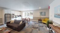 Located at 2445 S 222nd St in Des Moines, Washington, this single-level community clubhouse offers a well-appointed collection of shared amenity spaces designed to support everyday living and recreation. The welcoming entryway corridor features patterned carpet tiles in cool gray and earthy tones, setting a modern and cohesive aesthetic throughout the facility.

The central lounge and gathering area serves as the heart of the space, combining a comfortable seating arrangement with a dining area and an adjacent kitchenette outfitted with white cabinetry and appliances. Large windows overlook an outdoor pool area, filling the room with natural light and providing pleasant exterior views. Decorative accents, including nautical-themed artwork and vibrant accent chairs in bold yellow, lend a cheerful, curated character to the space.

The fitness room is equipped with a full dumbbell rack, resistance equipment, exercise mats, and a wall-mounted flat-screen television. Floor-to-ceiling mirrors amplify the sense of space and support workout visibility, while rubber flooring provides a practical and durable surface.

A dedicated laundry facility rounds out the amenities, housing multiple full-size washers and dryers in a clean, well-lit room with easy access and safety equipment conveniently mounted on the wall. Together, these spaces form a functional and inviting community hub.