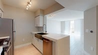 This single-floor residence presents a thoughtfully designed open-concept layout finished in a neutral, light-toned palette throughout. Light-format tile flooring extends across the living and kitchen areas, contributing to a bright and cohesive aesthetic.

The kitchen serves as a functional centerpiece, featuring white shaker-style cabinetry, a quartz waterfall peninsula, a deep stainless steel farmhouse sink with a professional-style pull-down faucet, and a full suite of stainless steel appliances including a range, refrigerator, and built-in microwave. A subway tile backsplash adds a subtle decorative detail. The peninsula opens directly to the living area, reinforcing the open-concept flow.

The living area is generously proportioned, with floor-to-ceiling vertical blinds framing expansive windows that allow abundant natural light and provide outdoor views. A secondary bedroom offers a bright, airy feel with a window and light-toned flooring.

The bathroom is finished with a white vanity, quartz countertop, large frameless mirror with bar lighting, and a soaking tub with tiled surround. In-unit laundry is conveniently housed behind bifold doors, accompanied by additional closet storage nearby.