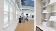 This single-story fitness center is designed to accommodate a comprehensive range of exercise activities across multiple clearly defined zones. The main workout area is characterized by its striking architectural detail — elevated tray ceilings painted in a bold blue contrast against crisp white walls and beams, creating a bright, energizing atmosphere. Ceiling fans are strategically placed throughout to maintain airflow. Large banks of windows along the perimeter walls flood the space with natural light while offering outdoor views, reinforcing an open, airy feel.

The primary exercise floor features neutral carpet and houses an extensive selection of equipment, including a row of treadmills, free weights, weight benches, a cable machine system, and various strength-training stations. Full-length mirrors are mounted along key walls to maximize both light and visibility during workouts. Built-in shelving near the entrance stores neatly folded towels for convenience.

A separate yoga and cycling studio is accessed through floor-to-ceiling glass sliding doors. This room features warm hardwood flooring, blue-painted walls, natural light from transom-style windows, and a dedicated spin bike. A direct exterior exit door provides additional accessibility and ventilation to the facility.