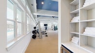 This single-story fitness center is designed to accommodate a comprehensive range of exercise activities across multiple clearly defined zones. The main workout area is characterized by its striking architectural detail — elevated tray ceilings painted in a bold blue contrast against crisp white walls and beams, creating a bright, energizing atmosphere. Ceiling fans are strategically placed throughout to maintain airflow. Large banks of windows along the perimeter walls flood the space with natural light while offering outdoor views, reinforcing an open, airy feel.

The primary exercise floor features neutral carpet and houses an extensive selection of equipment, including a row of treadmills, free weights, weight benches, a cable machine system, and various strength-training stations. Full-length mirrors are mounted along key walls to maximize both light and visibility during workouts. Built-in shelving near the entrance stores neatly folded towels for convenience.

A separate yoga and cycling studio is accessed through floor-to-ceiling glass sliding doors. This room features warm hardwood flooring, blue-painted walls, natural light from transom-style windows, and a dedicated spin bike. A direct exterior exit door provides additional accessibility and ventilation to the facility.
