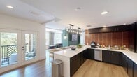 Located in Cary, NC, this thoughtfully designed single-floor community amenity space offers a generous footprint with a cohesive modern aesthetic throughout. Light-toned hardwood flooring runs continuously across the space, anchoring a clean, contemporary palette of crisp whites, deep teal accents, and dark wood paneling that adds warmth and visual contrast.

The main hallway serves as a welcoming spine, featuring a sputnik-style pendant chandelier, curated wall art, and a cozy seating vignette with mustard-yellow accent chairs — ideal for casual conversation or relaxed lounging. This corridor opens seamlessly into the various activity zones that define the space.

The games area is a standout feature, housing a full-size pool table with wood-plank accent walls and a wall-mounted television, alongside a separate shuffleboard room lined with large windows that flood the space with natural light and views of mature trees outdoors. An elegant bar-style kitchen island with dramatic dark cabinetry and pendant lighting provides a social hub, with counter seating for multiple guests.

The dining room offers two distinct seating configurations: a formal long-table setting beneath a striking multi-arm candle-style chandelier with garden views, and an intimate banquette nook framed by a textured accent wall with modern pendant lighting. Together, these spaces create a versatile, well-appointed environment suited for both social gatherings and private meetings.