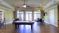 This single-floor recreational space is designed for leisure and social gathering, offering a well-appointed game room as its centerpiece. The room features rich hardwood flooring that extends throughout, lending warmth and a classic character to the space. A full-size pool table anchors the center of the room, providing a focal point for entertainment and friendly competition. Multiple ceiling fans are suspended overhead, complementing the recessed lighting and crown molding that frame the room with a refined, finished look. The walls are painted in a soft sage green tone, creating a relaxed and inviting atmosphere. Toward the back of the space, a dedicated seating area with round tables and upholstered chairs offers an ideal setting for card games or casual gathering. A wall-mounted flat-screen television adds a modern entertainment element to the room. Large French doors line the rear wall, flooding the interior with abundant natural light and providing direct access to an outdoor area. The overall ambiance strikes a balance between casual comfort and polished design, making this space well-suited for community recreation and social activity.