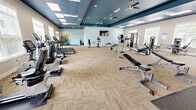 Located at 2300 Bluff Oak Way in Tallahassee, FL, this generously sized fitness center offers a comprehensive exercise environment across a single, open floor. The space is designed with a distinctive two-tone tray ceiling — painted a rich navy blue over neutral gray-beige walls — creating a polished, modern aesthetic that sets it apart from a typical gym setting. Multiple ceiling fans and recessed lighting, combined with a grid of skylight panels, ensure the space remains bright and comfortable throughout the day. Large windows along the perimeter walls flood the interior with natural light and provide views of the surrounding outdoor greenery.

The flooring features a combination of patterned carpet tiles in a neutral herringbone-style layout and rubber mat sections designated for free weight and high-impact activity areas. Equipment is thoughtfully organized throughout the space, with a dedicated cardio zone housing treadmills, ellipticals, and stationary bikes along the windowed wall. A strength training section includes cable machines, multi-functional weight stations, and benches, while a separate free weights area offers a full rack of kettlebells and dumbbells alongside wall-length mirrors. An additional open area accommodates functional fitness accessories including stability balls, medicine balls, and exercise mats. A wall-mounted television and water cooler further enhance the user experience.