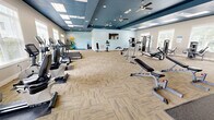 Located at 2300 Bluff Oak Way in Tallahassee, FL, this generously sized fitness center offers a comprehensive exercise environment across a single, open floor. The space is designed with a distinctive two-tone tray ceiling — painted a rich navy blue over neutral gray-beige walls — creating a polished, modern aesthetic that sets it apart from a typical gym setting. Multiple ceiling fans and recessed lighting, combined with a grid of skylight panels, ensure the space remains bright and comfortable throughout the day. Large windows along the perimeter walls flood the interior with natural light and provide views of the surrounding outdoor greenery.

The flooring features a combination of patterned carpet tiles in a neutral herringbone-style layout and rubber mat sections designated for free weight and high-impact activity areas. Equipment is thoughtfully organized throughout the space, with a dedicated cardio zone housing treadmills, ellipticals, and stationary bikes along the windowed wall. A strength training section includes cable machines, multi-functional weight stations, and benches, while a separate free weights area offers a full rack of kettlebells and dumbbells alongside wall-length mirrors. An additional open area accommodates functional fitness accessories including stability balls, medicine balls, and exercise mats. A wall-mounted television and water cooler further enhance the user experience.
