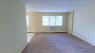 This single-floor space offers a clean, functional layout designed for comfortable everyday living. The main living area is generously proportioned, featuring wall-to-wall carpeting that adds warmth underfoot and a soft, neutral aesthetic throughout. Light-toned walls keep the interior feeling bright and open, while a large central window allows ample natural light to fill the room, offering views of the greenery outside. A secondary window on the adjacent wall further enhances the sense of airiness and openness. Climate control is well-considered, with a wall-mounted air conditioning unit positioned beneath the main window providing cooling, while baseboard heating runs along the perimeter of the room to ensure warmth during colder months. The smooth white ceiling completes the clean, uncluttered look of the space. Overall, the unit presents a straightforward and practical interior with a calm, neutral ambiance, making it well-suited for a variety of furniture arrangements and personal styles. The open feel of the living area, combined with its natural light and dual climate control systems, provides a solid foundation for a comfortable and versatile living environment.