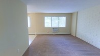 This single-floor space offers a clean, functional layout designed for comfortable everyday living. The main living area is generously proportioned, featuring wall-to-wall carpeting that adds warmth underfoot and a soft, neutral aesthetic throughout. Light-toned walls keep the interior feeling bright and open, while a large central window allows ample natural light to fill the room, offering views of the greenery outside. A secondary window on the adjacent wall further enhances the sense of airiness and openness. Climate control is well-considered, with a wall-mounted air conditioning unit positioned beneath the main window providing cooling, while baseboard heating runs along the perimeter of the room to ensure warmth during colder months. The smooth white ceiling completes the clean, uncluttered look of the space. Overall, the unit presents a straightforward and practical interior with a calm, neutral ambiance, making it well-suited for a variety of furniture arrangements and personal styles. The open feel of the living area, combined with its natural light and dual climate control systems, provides a solid foundation for a comfortable and versatile living environment.