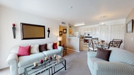 This single-floor residence offers a thoughtfully designed layout that maximizes functionality and comfort. The open-concept living and dining area serves as the heart of the home, featuring plush carpeting, neutral walls, and a sliding glass door that allows natural light to fill the space while providing access to the outdoors. A decorative wall mirror and wall-mounted sconces add character to the living area.

The galley-style kitchen is efficiently arranged with light wood cabinetry, laminate countertops, a double-basin sink, and white appliances including a refrigerator and electric range. Large tile flooring extends throughout the kitchen and connects seamlessly to a utility area housing the electrical panel.

The primary bedroom is generously sized, featuring carpet flooring, two windows dressed with bold red valances, and ample space for a full dresser set. A second bedroom mirrors the comfortable scale of the primary, complete with its own window, walk-in closet access, and carpeted floors — well-suited for use as a guest room or secondary sleeping space.

The bathroom is finished with large-format tile flooring, a single-sink vanity with under-cabinet storage, a well-lit mirror, and a separate toilet area. The overall design of the residence is clean and practical, offering a cohesive and livable floor plan throughout.