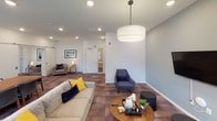 This single-floor community amenity space is thoughtfully designed to serve as a versatile gathering hub. The layout flows across several distinct functional zones, each offering a unique purpose while maintaining a cohesive contemporary aesthetic throughout.

The kitchen is a standout feature, outfitted with rich dark-wood cabinetry, stainless steel appliances including a full-size refrigerator and range, a white subway tile backsplash, granite countertops, and a central island with pendant lighting. Deep navy accent walls and warm hardwood-style flooring lend the kitchen area a bold, inviting character. A built-in bench with a bright yellow cushion adds a casual seating nook nearby.

The lounge areas transition into softer tones, with walls in light gray-blue and plush patterned carpet flooring underfoot. A large sectional sofa and coordinating armchairs in navy and gray are anchored by a round wood coffee table, with a wall-mounted television making it ideal for relaxed gatherings. Recessed lighting and a large drum pendant fixture provide even, welcoming illumination. Access to an outdoor area is available through a glass door, allowing natural light to filter in.

Additional seating nooks, a billiards table, a dining table with ample seating, and high-top bar seating round out the amenities, making this a comprehensive and functional shared space.