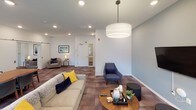This single-floor community amenity space is thoughtfully designed to serve as a versatile gathering hub. The layout flows across several distinct functional zones, each offering a unique purpose while maintaining a cohesive contemporary aesthetic throughout.

The kitchen is a standout feature, outfitted with rich dark-wood cabinetry, stainless steel appliances including a full-size refrigerator and range, a white subway tile backsplash, granite countertops, and a central island with pendant lighting. Deep navy accent walls and warm hardwood-style flooring lend the kitchen area a bold, inviting character. A built-in bench with a bright yellow cushion adds a casual seating nook nearby.

The lounge areas transition into softer tones, with walls in light gray-blue and plush patterned carpet flooring underfoot. A large sectional sofa and coordinating armchairs in navy and gray are anchored by a round wood coffee table, with a wall-mounted television making it ideal for relaxed gatherings. Recessed lighting and a large drum pendant fixture provide even, welcoming illumination. Access to an outdoor area is available through a glass door, allowing natural light to filter in.

Additional seating nooks, a billiards table, a dining table with ample seating, and high-top bar seating round out the amenities, making this a comprehensive and functional shared space.