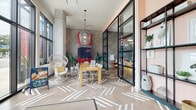 This single-floor space makes an immediate impression with its energetic, design-forward aesthetic and welcoming atmosphere. Large, floor-to-ceiling windows line the exterior walls, flooding the interior with abundant natural light and offering open views of the surrounding streetscape. Exposed concrete columns and polished concrete floors featuring bold white geometric patterns add an industrial-chic character throughout.

The space is thoughtfully curated with an eclectic mix of seating, including woven rattan peacock chairs, leather safari-style chairs, and mixed-material dining chairs arranged around a wooden table — creating distinct, inviting seating areas. A striking feature wall clad in diagonal wood-tone paneling serves as a dramatic backdrop, accented by a bold red "See You Later Alligator" wall art installation that adds a playful, branded identity to the space.

A steel-framed glass partition wall with black mullions separates areas while maintaining visual openness, and a tall black open-shelf display unit against a soft blush-pink wall showcases curated artwork, plants, and decorative objects. Woven pendant lights add warmth overhead, complementing the lush potted plants that are scattered throughout. The overall ambiance is modern, artistic, and vibrant — designed to engage and inspire visitors from the moment they arrive.