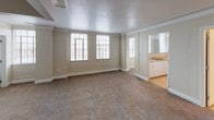 This single-floor unit offers a well-distributed layout with multiple rooms connected by a central corridor, as seen in the 3D floor plan. Throughout the living and bedroom areas, warm beige carpet flows consistently, complemented by crisp white crown molding and neutral-toned walls that create a clean, understated aesthetic.

The generously sized living areas provide ample room for furnishings, with several spaces featuring gridded windows that allow natural light to filter in. Closets with built-in rod systems are present in multiple rooms, offering practical storage solutions. Crown molding details are a recurring architectural feature, lending a refined character to each carpeted space.

The kitchen is equipped with white cabinetry, a coil-burner electric range, and a refrigerator, with tile flooring that transitions from the surrounding carpet. A mirrored vanity and sink area is positioned adjacent to one of the larger rooms, adding convenience.

The bathroom features light tile flooring extending into a hallway-style layout, with a toilet and grab bar visible, suggesting thoughtful accessibility considerations. Overall, the unit maintains a neutral, move-in-ready palette with consistent finishes throughout.