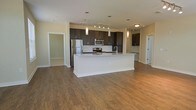 This single-floor residence showcases a thoughtfully designed open-concept layout that seamlessly connects the kitchen and living areas. The kitchen serves as the centerpiece, anchored by a large white island with a light stone countertop that provides generous workspace and casual seating potential. Dark espresso-toned cabinetry lines the walls, offering abundant storage while creating a bold contrast against the neutral, warm-toned walls. Stainless steel appliances, including a refrigerator, microwave, and range, complement the contemporary aesthetic. Pendant lighting hangs above the island, while a track lighting system illuminates the cabinetry and cooking area. Hardwood-style flooring flows throughout the open space, adding warmth and continuity to the overall design. The expansive layout of the main living area allows for flexible furniture arrangements. Multiple doorways visible from the main space suggest additional rooms, each framed by clean white trim that adds a crisp, finished look. The overall ambiance is modern and functional, with a neutral color palette and quality finishes that create a clean, inviting atmosphere.