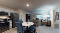Located at 1525 W Arrowood Rd in Charlotte, NC, this single-floor suite offers a thoughtfully designed layout ideal for extended stays or comfortable living. The open-concept living and dining area is anchored by light wood-style flooring throughout, a round dining table with four blue upholstered chairs, and a cozy living room furnished with neutral-toned sofas and accent artwork. A flat-screen TV and media console complete the lounge area, which benefits from natural light through large windows with blinds.

The fully equipped kitchen features white cabinetry with granite-style countertops, stainless steel appliances including a refrigerator, range, microwave, and dishwasher — offering everything needed for home-style cooking. Each of the two bedrooms provides a comfortable retreat, with king-size beds, coordinating artwork, wall-mounted televisions, and open wire-rack closets offering ample storage space. Both bedrooms are brightened by windows with curtains.

The suite includes two bathrooms: one with a glass-enclosed shower and another with a full soaking tub and shower combination. Both bathrooms feature granite-topped white vanities and wood-look flooring. A stacked washer and dryer is also included within the unit, adding everyday convenience to this well-rounded Charlotte suite.