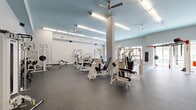 This expansive fitness center occupies a single, open floor plan designed to accommodate a wide range of workout disciplines. The space is characterized by its generous square footage, allowing for a thoughtful distribution of equipment zones without feeling crowded. Durable rubber flooring covers the entire area, providing safety and sound absorption throughout.

The strength training section features multiple Cybex selectorized machines, a squat rack with plate-loaded barbells, adjustable benches, and a comprehensive free weight area with organized dumbbell racks and medicine balls. A dedicated functional training zone includes a bold red multi-station rig equipped with a heavy BOX punching bag, gymnastic rings, TRX-style suspension straps, and resistance bands, catering to dynamic and bodyweight training.

The cardio area houses a row of treadmills and elliptical machines positioned along the perimeter. Elevated ceilings with blue accent details add architectural interest, while ceiling fans and fluorescent lighting ensure a well-ventilated, brightly lit environment. Large windows along one wall invite natural light and provide outdoor views. A wall-mounted TV, whiteboard, and storage cubbies add functional amenities. The overall ambiance is clean, modern, and professional.