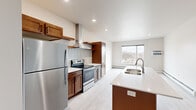 This well-designed 2-bedroom, 1-bathroom unit offers a thoughtfully arranged single-floor layout with contemporary finishes throughout. Light wood-look flooring runs consistently across all rooms, creating a cohesive and clean aesthetic.

The kitchen serves as a central highlight, featuring warm-toned wood cabinetry, stainless steel appliances including a refrigerator and electric range, a stainless wall-mounted range hood, and a herringbone tile backsplash. A kitchen island with a white quartz countertop and undermount double sink provides additional workspace and separates the kitchen from the open living area, which receives natural light through a large window.

The primary bedroom is generously proportioned and includes a closet with bifold doors and baseboard heating. Its layout comfortably accommodates standard furnishings. The second bedroom features a large picture window alongside clerestory windows, offering broad outdoor views and ample natural light.

The bathroom includes a wood-toned vanity with a white countertop, an undermount sink, and a large mirror, finished with the same light flooring found throughout the unit. The overall design is clean and modern, with neutral tones and consistent materials providing a move-in-ready environment.
