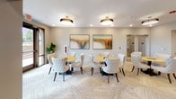 Located at 5735 Dixie River Road in Charlotte, NC, this single-floor community amenity space is thoughtfully designed to offer both social and recreational experiences. The interior is finished with a neutral palette of soft grays and warm beiges, complemented by patterned herringbone carpeting that flows throughout the space, lending a sense of cohesion and understated elegance.

The dining and lounge area features multiple round wooden tables surrounded by tufted, upholstered chairs in a light cream tone, creating an inviting setting for casual gatherings. Overhead, stylish drum pendant light fixtures provide warm, even illumination, while framed abstract landscape artwork adds vibrant color to the walls. Natural light filters in through large windows and glass-paneled doors, connecting the interior to the outdoors.

Transitioning deeper into the space, the game room area makes a bold design statement with a rich teal accent wall. A full-sized pool table anchors this zone, while a large wall-mounted Scrabble board serves as both a decorative and interactive feature. Deep navy velvet lounge chairs and a sleek side table create a comfortable seating vignette nearby. Recessed lighting and pendant fixtures ensure consistent brightness throughout, making this a well-appointed, versatile communal retreat.