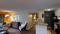 This single-story residence is designed with an open-concept floor plan that promotes a natural flow between the main living spaces. The living area is comfortably proportioned, featuring rich hardwood-style flooring that extends throughout the home, adding warmth and visual continuity. A textured ceiling runs across the space, complemented by neutral-toned walls that provide a versatile backdrop for various décor styles.

The kitchen is positioned adjacent to the living area, separated by a half-wall peninsula that doubles as a breakfast bar. It features white cabinetry, a single-basin sink, and light countertops, keeping the space bright and functional. The peninsula design enhances the open feel while maintaining a defined cooking zone.

A corridor visible from the main living area leads to additional rooms, including what appears to be a bedroom, suggesting a thoughtful layout that separates private and shared spaces. The overall ambiance is relaxed and inviting, with consistent flooring and a cohesive neutral color palette tying all areas together into a unified, comfortable living environment.