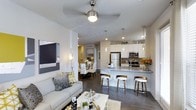 This well-appointed single-floor residence offers a seamless open-concept layout that connects the living, dining, and kitchen areas into one cohesive and functional space. The living room is anchored by a plush gray sofa, a ceiling fan with integrated lighting, and a contemporary art piece that adds character to the bright white walls. Dark hardwood flooring runs throughout, providing a striking contrast to the light-toned furnishings and cabinetry. The kitchen is a standout feature, boasting crisp white cabinetry that extends to the ceiling, stainless steel appliances — including a refrigerator, microwave, and range — and a granite-topped peninsula that doubles as a breakfast bar with stylish bar seating. Elegant pendant lights hang above the peninsula, complementing the recessed lighting that keeps the space bright and inviting. Adjacent to the kitchen, a dedicated dining area accommodates a full table and seating, enhanced by a wall-mounted wine rack that adds a refined touch. The overall ambiance is modern and sophisticated, with a neutral color palette punctuated by warm accent tones, creating a space that feels both polished and comfortable.