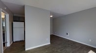 This single-floor space offers a clean, modern aesthetic defined by a thoughtfully open layout. The interior is finished with dark hardwood-style flooring that runs throughout, providing a warm contrast against the cool, neutral gray walls. Bright white baseboards frame the floors, adding a crisp, polished detail to each room. The living area is open and versatile, with a flush-mount ceiling light providing functional illumination. A wall-mounted heating unit is conveniently integrated into the living space. The textured white ceiling adds subtle visual interest overhead. The space transitions smoothly into the kitchen area, where a full-size white refrigerator is visible, hinting at a practical and functional kitchen layout. A partial wall divides the kitchen from the main living area, creating a sense of definition without sacrificing the open feel of the overall floor plan. The neutral color palette throughout creates a bright, airy ambiance that lends itself well to a variety of interior styles, making this a flexible and inviting space.