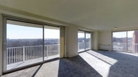 This single-floor space is defined by its generous, open living area, designed to maximize natural light and outdoor views. Two sets of large sliding glass doors span much of the exterior wall, flooding the interior with sunlight and providing direct access to a railed balcony. The elevated position of the unit affords broad, unobstructed panoramic views of the surrounding urban landscape and tree canopy, visible through the full-width glazing.

The living area features neutral-toned carpeting that transitions to a lighter, tile-like flooring section near the balcony doors, creating a subtle distinction within the open layout. The walls are finished in a clean off-white, complementing the smooth, flat ceiling that runs throughout the space. Vertical blinds are installed along the larger sliding door, offering adjustable light and privacy control.

A baseboard heating unit is positioned along one wall, indicating a straightforward climate system. The overall ambiance is clean, airy, and minimalist, with the expansive windows and balcony access serving as the defining architectural features of this light-filled space.