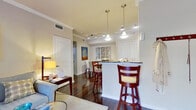 This single-story residence offers a thoughtfully designed layout with a cohesive blend of warm and neutral tones throughout. Rich, dark hardwood flooring flows through the main living areas, creating a polished and inviting atmosphere accentuated by crown molding and clean white trim.

The kitchen is well-equipped with warm cherry wood cabinetry, dark granite countertops, a mosaic tile backsplash, and stainless steel appliances including a range, microwave, and refrigerator. A breakfast bar with bar seating connects the kitchen to the main living space, offering a casual dining option.

The open-concept layout accommodates both a living room and a dedicated dining area. The living room features a comfortable sectional sofa and a glass-top coffee table, while the dining area hosts a round wood table with upholstered chairs and decorative wall art. A built-in desk nook provides a functional workspace with shelving and natural light from a nearby door.

The bedroom is carpeted and serene, featuring a large window with garden views, elegant drapery, and access to a spacious walk-in closet. Crown molding continues into this room, reinforcing the refined character found throughout the residence.