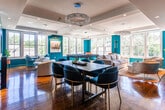 Located at 31 Vose Ave in South Orange, NJ, this single-floor space makes a bold design statement with its signature teal accent walls and warm hardwood flooring throughout. The open layout flows seamlessly between distinct functional areas, creating an inviting and energetic atmosphere.

The main lounge area is anchored by a striking teal feature wall housing a wall-mounted television, complemented by an elegant crystal chandelier overhead. Built-in teal bookshelves add both visual interest and practical storage. Abundant natural light pours in through large double-hung windows, offering pleasant views of the surrounding greenery.

A dedicated recreation zone centers around a sleek black billiards table, set beneath a coffered ceiling with a decorative chandelier. Multiple oversized windows wrap this area, flooding it with daylight and creating an airy, expansive feel.

The kitchenette is well-appointed with rich dark-wood cabinetry, granite countertops, a mosaic tile backsplash, and stainless steel appliances. Adjacent to it, a dining area with blue velvet chairs and a dark table provides a stylish spot for gathering.

A cozy nook near the entrance features a teal media wall with a built-in television and an intimate café-style seating arrangement. The entry corridor leads visitors in with white paneled doors and refined finishes, setting the tone for this thoughtfully designed, vibrant space.