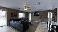 This single-storey space centres around a well-appointed living area designed for comfort and functionality. The room is finished with neutral taupe-toned walls that create a warm, grounded atmosphere, complemented by a combination of tile and carpeted flooring that defines the living zone. A large sectional sofa in dark leather anchors the seating area, paired with a low-profile coffee table positioned atop an area rug. A wall-mounted flat-screen television serves as a focal point on one wall, while a decorative vase with dried botanicals adds a subtle organic touch to the space. Natural light enters through a window overlooking the exterior, brightening the room during daylight hours. The ceiling features a wood-bladed ceiling fan with an integrated light fixture, supported by additional flush-mount light fixtures distributed throughout. A textured ceiling adds a classic finishing detail. The overall ambiance is relaxed and practical, with a layout that balances open circulation space with defined areas for lounging and entertainment.