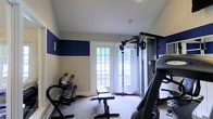 Located at 5450 Firewater Ln in Westerville, Ohio, this single-floor home gym is thoughtfully designed to support a well-rounded fitness routine. The space is defined by striking navy blue accent bands along the upper walls, contrasting cleanly with the neutral lower walls and light ceiling to create an energizing yet focused atmosphere. Large, white-framed French doors flood the room with natural light and offer a pleasant view of the surrounding greenery, making workouts feel open and inviting rather than confined. The carpeted flooring provides a practical surface underfoot throughout the space. The room is equipped with a full multi-station cable and pulley weight machine ideal for a wide range of strength training exercises, a rack of graduated dumbbells for versatile free weight training, a flat workout bench, and a stationary cycling bike for cardiovascular conditioning. Recessed ceiling speakers add an audio element to enhance the workout environment. The combination of functional equipment, ample natural light, and bold design choices makes this home gym a practical and motivating space for daily fitness use.