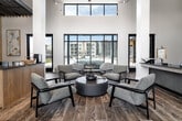 This generously sized, single-floor amenity space is thoughtfully designed to accommodate a variety of social and professional activities. The entrance opens into a dramatic reception lounge defined by soaring double-height ceilings, bold structural columns, and floor-to-ceiling windows that frame outdoor views of a pool area, flooding the interior with natural light. Track lighting above complements the bright, airy atmosphere throughout.

The main lounge area features multiple distinct seating arrangements, including a central conversation cluster with mid-century modern chairs around a round coffee table, as well as a more relaxed living area anchored by a sectional sofa, an accent chair, and a wall-mounted fireplace. A billiards table and dedicated workspace desks are visible in adjoining zones, catering to both leisure and productivity.

A fully equipped kitchenette provides light cabinetry in warm wood tones, a striking diagonal-tile backsplash in slate blue, open shelving, a farmhouse sink, stainless steel appliances, and a marble-topped island with bar seating. Textured accent walls in neutral tones add depth to breakout seating areas with a vibrant blue sectional sofa and private glass-enclosed meeting rooms with interior windows. Wood-look tile flooring flows seamlessly throughout, tying the entire space together cohesively.