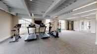 Located at 77 Water St in Warren, Rhode Island, this fitness center offers a thoughtfully designed, single-floor workout space that balances functionality with a clean, modern aesthetic. The exposed structural ceiling, painted in light gray, lends an industrial character to the room while maintaining an open, airy atmosphere. Linear LED lighting runs along the ceiling framework, ensuring consistent illumination throughout the space.

Large windows along the perimeter walls flood the interior with natural light and provide views of the surrounding outdoor environment, enhancing the overall workout experience. The floor is finished with durable, patterned carpet tile in neutral tones, providing comfort underfoot across the expansive workout area.

The cardio zone features multiple treadmills and an elliptical trainer, complemented by a wall-mounted flat-screen television. A dedicated strength training area includes a multi-function cable machine, a dumbbell rack with a full range of weights, and a flat weight bench. Additional functional fitness accessories—including resistance bands, medicine balls, and yoga mats—are neatly organized on a freestanding storage rack. A mirrored wall section enhances the sense of space. Glass doors connect to an adjacent room, suggesting additional amenity spaces nearby.
