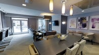 This refined single-floor space seamlessly blends lounge and social gathering functions within a thoughtfully designed interior. The open layout features a large dark dining or conference table at the forefront, surrounded by plush, nailhead-trimmed upholstered chairs in a warm neutral tone, setting an inviting yet polished atmosphere.

Beyond the table, a comfortable lounge area unfolds, furnished with a generous sofa and accent chairs arranged around a sleek coffee table. A textured stone accent wall serves as a striking focal point, anchoring a wall-mounted flat-screen television flanked by stylish wall sconces that cast a warm, ambient glow.

The ceiling is detailed with tiered recesses and fitted with a mix of cylindrical pendant lights and recessed fixtures, contributing to a layered lighting scheme that enhances the space's contemporary character. Soft grey carpeting and warm wood-toned flooring elements add depth and comfort underfoot.

Large windows and a glass door at the far end of the room flood the interior with natural light and provide direct access to an outdoor terrace or patio, extending the usable space beyond its interior walls. Decorative artwork and carefully curated furnishings throughout reinforce the space's upscale, welcoming ambiance — well-suited for both casual socializing and more formal gatherings.