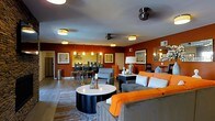 This single-floor residence showcases a thoughtfully designed open-concept layout that seamlessly connects the living, dining, and kitchen areas. The living room is anchored by a sleek, cylindrical coffee table in black and white, surrounded by plush seating in neutral tones accented with vibrant orange throw pillows and blankets, creating an inviting, contemporary atmosphere.

The bold terracotta-red accent walls provide a striking visual contrast against the light gray ceiling, which features multiple flush-mount and pendant light fixtures that ensure the space is well-lit throughout. A ceiling fan adds both function and style overhead.

The dining area, visible from the living room, includes a compact table with bar-height seating, illuminated by elegant pendant lights. Adjacent to it, the kitchen offers stainless steel appliances and under-cabinet lighting, contributing to the cohesive modern aesthetic.

A mirror on one wall reflects light and adds visual depth to the space. Decorative framed artwork adorns the walls, complementing the warm color palette. A large area rug defines the living zone, tying together the furnishings and adding texture to the hardwood-style flooring beneath.