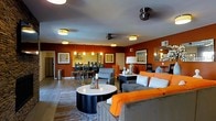 This single-floor residence showcases a thoughtfully designed open-concept layout that seamlessly connects the living, dining, and kitchen areas. The living room is anchored by a sleek, cylindrical coffee table in black and white, surrounded by plush seating in neutral tones accented with vibrant orange throw pillows and blankets, creating an inviting, contemporary atmosphere.

The bold terracotta-red accent walls provide a striking visual contrast against the light gray ceiling, which features multiple flush-mount and pendant light fixtures that ensure the space is well-lit throughout. A ceiling fan adds both function and style overhead.

The dining area, visible from the living room, includes a compact table with bar-height seating, illuminated by elegant pendant lights. Adjacent to it, the kitchen offers stainless steel appliances and under-cabinet lighting, contributing to the cohesive modern aesthetic.

A mirror on one wall reflects light and adds visual depth to the space. Decorative framed artwork adorns the walls, complementing the warm color palette. A large area rug defines the living zone, tying together the furnishings and adding texture to the hardwood-style flooring beneath.