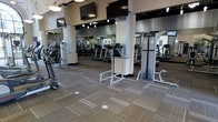 This fitness center offers a comprehensive and well-organized workout environment designed to accommodate a variety of exercise routines. The space is outfitted with an impressive selection of cardio equipment, including treadmills and elliptical machines, alongside dedicated strength training stations that cater to users of all fitness levels. Large wall-to-wall mirrors line the perimeter, enhancing the sense of openness and allowing users to monitor their form during workouts. Multiple flat-screen televisions are mounted at strategic heights throughout the space, ensuring visibility from virtually any machine or station. The flooring consists of durable, patterned carpet tiles that provide both comfort and a clean, modern aesthetic. Overhead, a combination of recessed lighting and pendant fixtures delivers bright, even illumination across the entire floor. The neutral color palette of warm beiges and deep accent tones creates a focused yet welcoming atmosphere. The layout is thoughtfully arranged to allow ample space between equipment, promoting ease of movement and a comfortable workout experience. Overall, this fitness center presents a polished, fully functional facility suited for daily use.