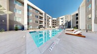 Located at 5050 W Russell Rd in Las Vegas, NV, this contemporary residential community showcases a bold modern architectural design with a contrasting exterior palette of dark gray, white, and terracotta-orange stucco facades. The multi-story building features private balconies, large windows, and clean geometric lines that frame a generously sized central courtyard.

At the heart of the outdoor amenity space is a expansive resort-style swimming pool with crystal-clear blue water, flanked by a dedicated sundeck lined with wood-framed chaise lounges. Adjacent to the pool, a separate spa provides an additional relaxation area, also surrounded by lounge seating and open concrete decking. Covered breezeway passages with recessed lighting connect the courtyard to building entrances, featuring glass panel railings and low-profile bollard lighting.

The landscaping throughout the courtyard reflects a desert-conscious design, incorporating decorative river rock, drought-tolerant plantings, native shrubs, and mature palm trees. Lush turf areas provide a visual contrast to the stone groundcover. Concrete pathways guide residents through the grounds toward building entry points framed by sleek vertical metal gates. The overall ambiance is clean, open, and well-appointed, offering a functional and attractive outdoor living environment suited to the Las Vegas climate.