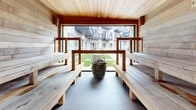 Located at 2274 Princess Street in Kingston, Ontario, this thoughtfully designed sauna offers a refined wellness retreat finished entirely in natural wood. The interior is clad in light, horizontal wood planking across the walls, ceiling, and tiered benches, creating a warm, cohesive aesthetic that emphasizes craftsmanship and comfort. Two rows of generously sized wooden benches line both sides of the sauna, providing ample seating at varying heights to accommodate different heat preferences. The slate-tiled floor anchors the space with a cool, contrasting tone that complements the warmth of the surrounding wood. A traditional stone sauna heater sits centrally at the far end, serving as both a functional and visual focal point. Natural light floods the interior through a large picture window and a glass-paneled door, both of which frame a view of a manicured green courtyard and the surrounding residential building. The window placement not only brightens the space but also creates a seamless visual connection to the outdoors, enhancing the sense of openness. The patio area adjacent to the sauna extends the experience outdoors, offering a natural transition space. Overall, this sauna combines classic Finnish sauna design with modern finishes to deliver a serene and functional wellness environment.