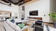 This single-floor space is a well-appointed amenity lounge designed with a striking contemporary aesthetic. The main living area commands attention with dramatically high ceilings featuring exposed dark ductwork and recessed lighting against a contrasting dark ceiling plane. Crisp white walls serve as a backdrop for curated abstract wall art, while a sleek, wall-mounted flat-screen television is anchored above a linear gas fireplace set into a minimalist surround, creating a refined focal point. Generous sectional sofas in light gray are accented with bold green-patterned and faux fur throw pillows, complemented by angular dark leather armchairs arranged around a low-profile wood coffee table.

The open layout flows into a game and dining zone, where a full-size billiards table is positioned near floor-to-ceiling windows that invite abundant natural light. A bold black-and-white geometric wallcovering defines the dining area, where a formal dining table sits beneath elegant pendant chandeliers. A compact kitchen includes stainless steel appliances, white cabinetry, quartz countertops, and a shimmering mosaic tile backsplash with a stainless range hood. The overall ambiance is sophisticated, energetic, and purposefully designed for social engagement.