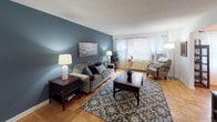 This single-floor residence offers a thoughtfully designed layout connecting a series of comfortable, functional spaces unified by a consistent design palette. Throughout the home, warm parquet flooring and cool blue accent walls create a cohesive and inviting atmosphere.

The living room is a standout space, anchored by a bold slate-blue feature wall adorned with landscape artwork. Natural light streams in through large windows dressed with sheer curtains, brightening the parquet floors and adding to the room's open, airy feel.

Adjacent to the living room, the dining area continues the blue accent wall theme and features an elegant chandelier overhead, a window with natural light, and ample space for a full dining set — ideal for everyday meals or entertaining.

The kitchen is well-equipped with light wood cabinetry, white appliances including a gas range and dishwasher, and the added convenience of an in-unit stacked washer and dryer. Durable tile flooring completes the space.

The bedroom is generously sized, featuring parquet flooring, a walk-in closet, and a window that brings in natural light. The room's soft blue-grey walls carry the home's design theme seamlessly.

The bathroom is clean and functional, offering a vanity with under-cabinet storage, a mirrored medicine cabinet, and a shower/tub combination.