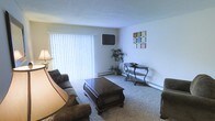 This single-floor residence offers a welcoming living room as its central gathering space. The room is finished with wall-to-wall carpeting in a neutral tone, providing a soft and comfortable underfoot feel that contributes to the overall warmth of the space. A large sliding glass door dressed with vertical blinds serves as the primary source of natural light, brightening the room while offering the option for privacy. The door also provides access to the outdoors, enhancing the sense of openness.

The living room is thoughtfully arranged with a dark upholstered sofa and a matching accent chair, centered around a rich, dark wood coffee table with a classic curved design. A sleek wall-mounted flat-screen television is positioned above baseboard heating, making it both practical and space-efficient. An elegant wrought-iron console table against the far wall adds a decorative touch, complemented by a small potted plant that introduces a natural element. Decorative wall art provides a pop of color against the neutral walls. The textured ceiling and clean lines throughout give the space a straightforward, functional aesthetic suited for comfortable day-to-day living.