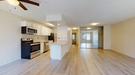 This single-floor residence is designed with a practical, open-concept layout that connects the kitchen and living areas seamlessly. The kitchen is well-equipped with stainless steel appliances, including a range, microwave, dishwasher, and refrigerator, complemented by white cabinetry, a subtle tile backsplash, and light-toned countertops. A central kitchen island provides additional workspace and serves as a natural divider between the kitchen and the open living area. Light hardwood-style flooring runs throughout the main living spaces, contributing to a bright, cohesive aesthetic.

Adjacent to the main living area, a bedroom features mirrored sliding closet doors, maximizing the sense of space. Additional bedrooms are carpeted and each include windows that invite natural light and outdoor views of surrounding greenery. The neutral wall tones and textured ceilings carry throughout the bedrooms, maintaining a consistent, understated style.

The bathroom is finished with white cabinetry featuring brushed nickel hardware, a stone-toned countertop, a large vanity mirror, and a full tub-shower combination with white tile surround. The light flooring in the bathroom coordinates well with the rest of the residence, creating a unified and clean overall ambiance.